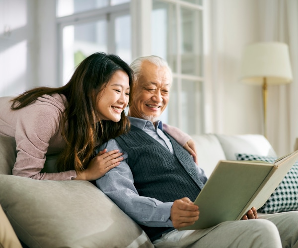 Diverse old man sitting on couch reading a book with his daughter reading over his shoulder